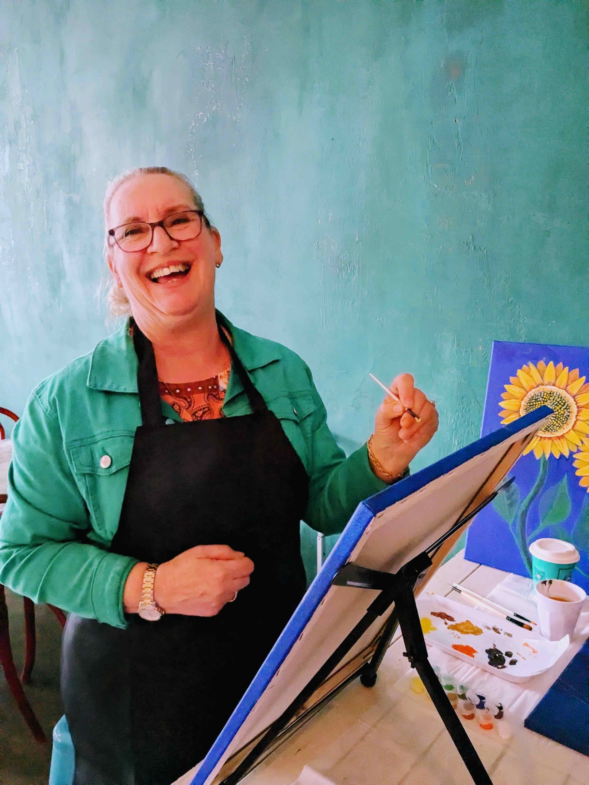 Artist in green jacket and black apron smiling while holding paintbrush at easel with sunflower painting
