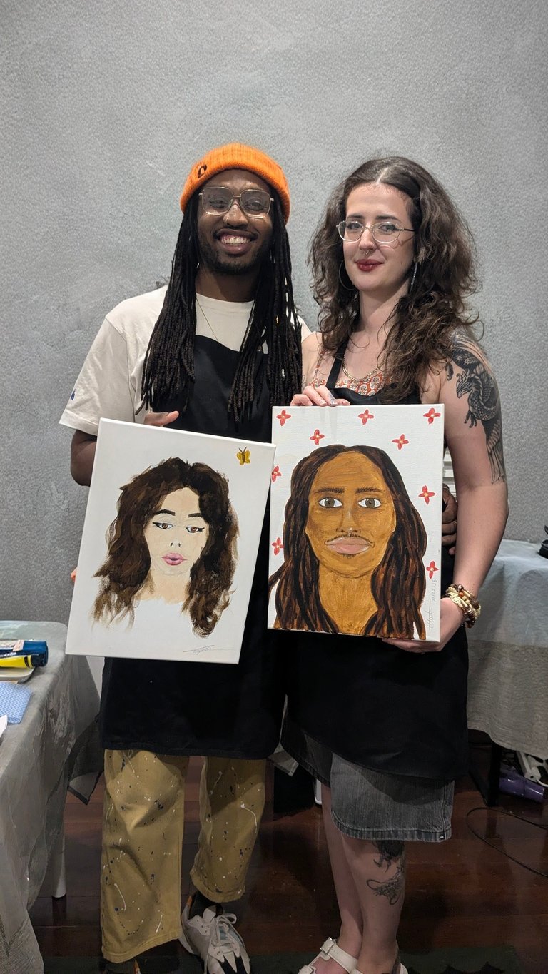 Two people smiling while holding portrait paintings against a gray wall