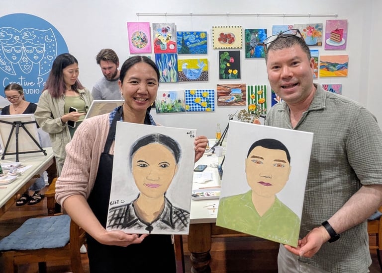 Woman and man holding portrait paintings in an art classroom with students and colorful artwork displayed on walls