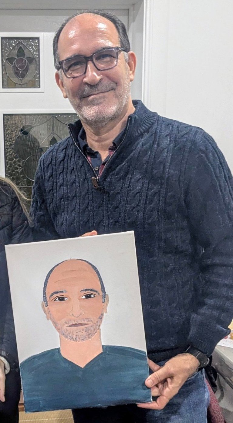 Man with glasses wearing a dark quilted jacket holds a portrait painting of a bald man with beard